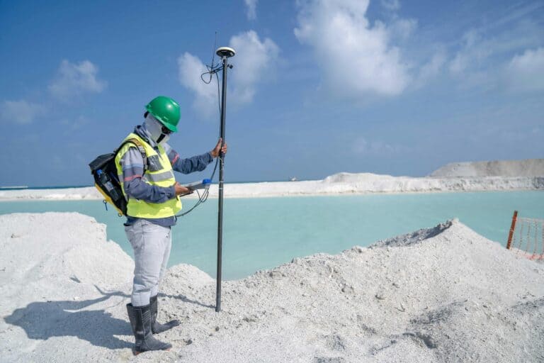 A surveyor wearing protective gear and a green hard hat uses GPS equipment to measure terrain near a light blue water basin during a bathymetric or land survey.
