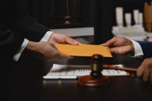 Close-up of two professionals exchanging a sealed envelope across a desk, with a judge’s gavel and legal documents in the foreground, suggesting a legal or ethical transaction scenario.
