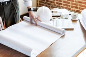 Person reviewing large architectural blueprints on a desk with a hard hat, rolled plans, and lighting components nearby in a modern office setting
