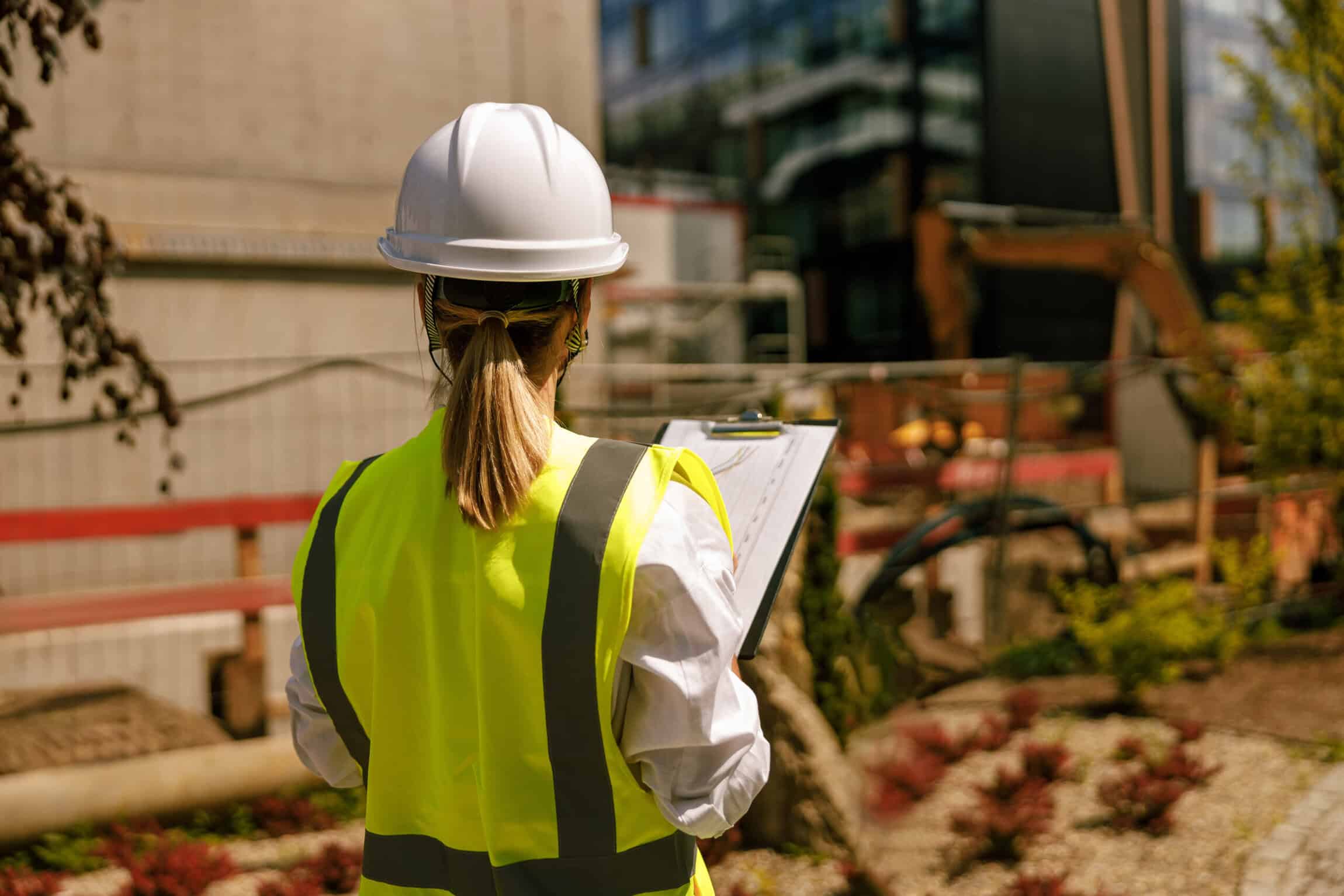 Construction worker in a hard hat and safety vest reviewing plans at an active construction site.